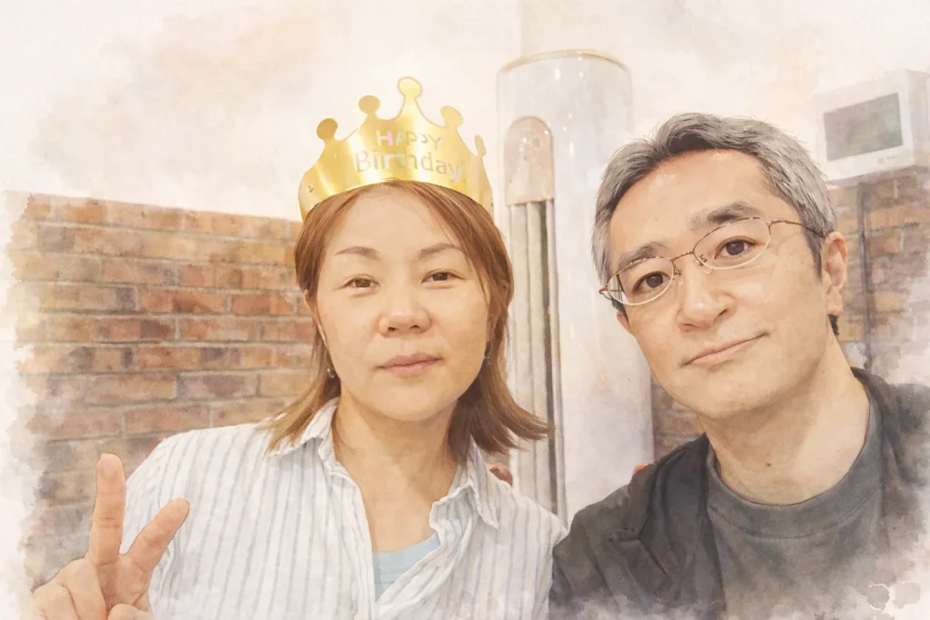 誕生日の「Happy Birthday」と書かれた金の王冠を被り、ピースサインをする中国人妻と、その隣で微笑む眼鏡をかけた日本人夫（運営者）のツーショット。水彩画のような柔らかなタッチの加工が施されており、温かみのある夫婦の日常の一コマを表現している。