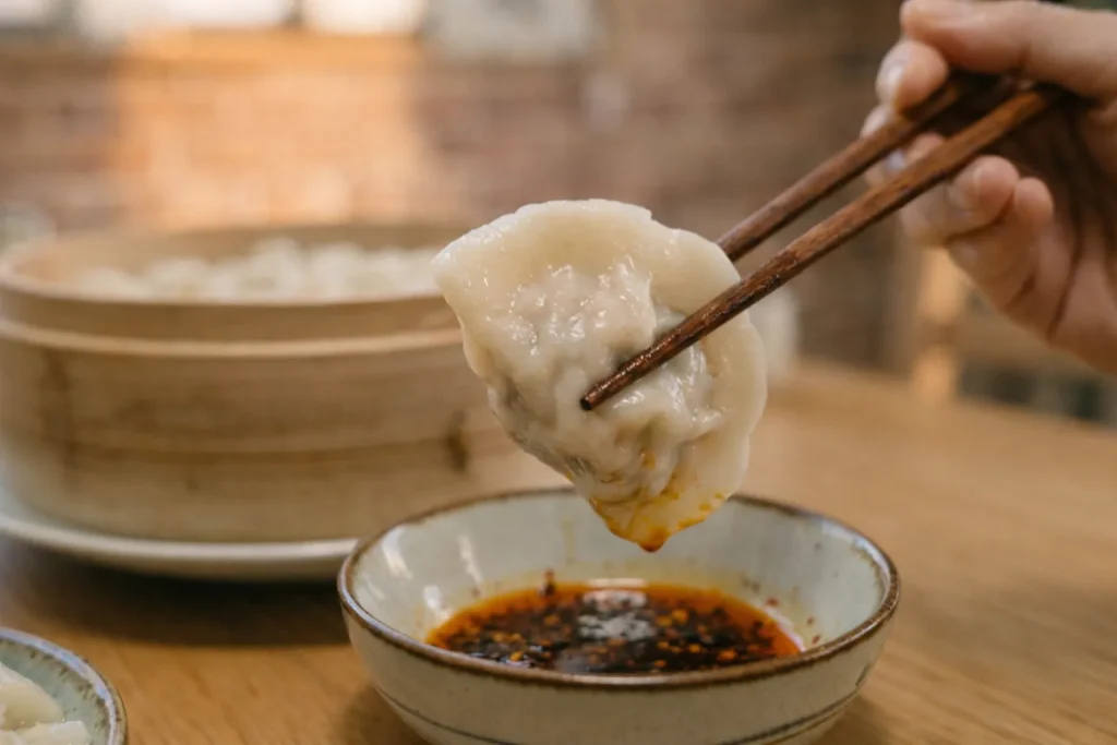 箸で水餃子を持ち上げ、ラー油入りのタレにつける瞬間のクローズアップ。中国の餃子の食べ方を表現した画像。